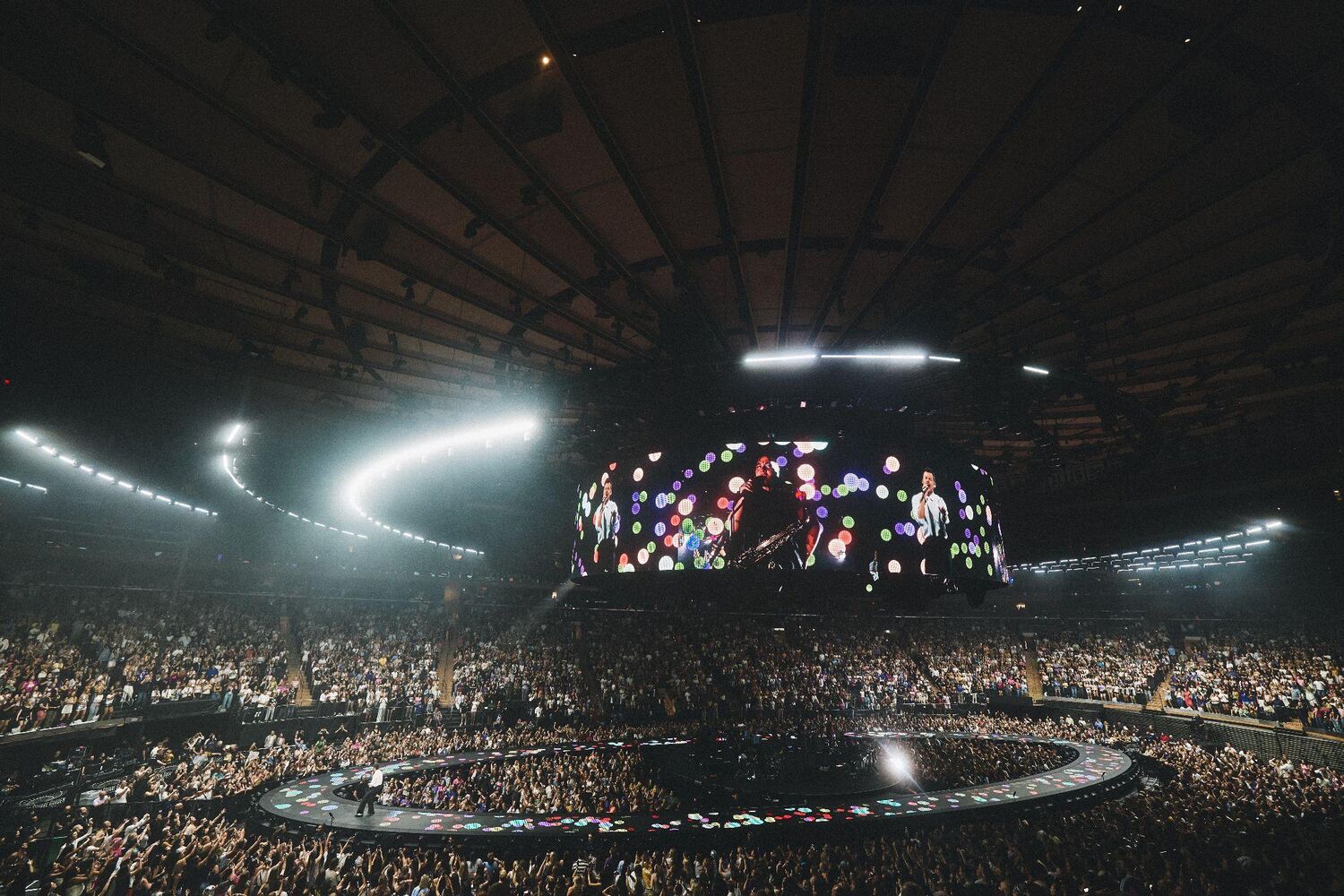 Inside a packed Madison Square Garden during Harry Styles' show, with arcs of white light under the ceiling and a giant video screen hung above a ring-shaped stage on the floor.